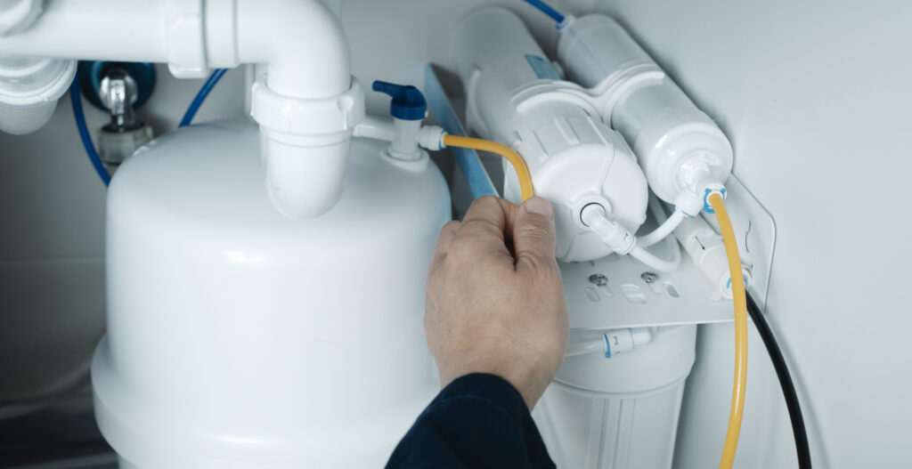 A close view of a plumber's hands as they install a reverse osmosis system under the sink in a kitchen.